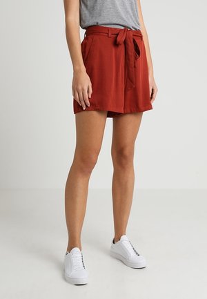 Shorts rouges en tissu léger avec une taille ceinturée, dotés de deux poches latérales et d'une coupe décontractée, associés à des baskets blanches.