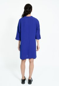 Ohlapna, obleka do kolen iz živahnega modrega materiala z okroglim izrezom in kratkimi rokavi. Zadrga na hrbtu, gladka tekstura. Črne čevlje.
