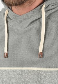Hoodie gris en maille avec un tissu texturé, cordons de serrage crème et accents en cuir marron aux extrémités des cordons.