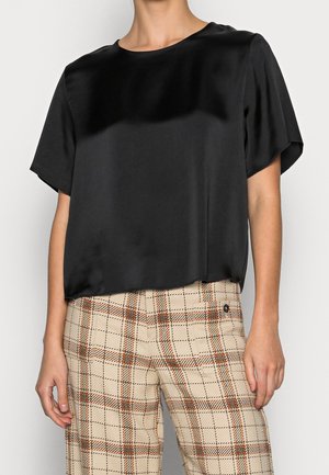 Person trägt eine kurzärmelige schwarze Satinbluse und beige karierte Hose mit orangefarbenen und dunkelbraunen Linien.