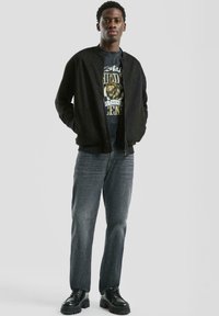Jonge man staat met zijn handen in de zakken van een zwarte bomberjack, draagt een t-shirt met een leeuwgraphic, grijze jeans en zwarte chunky schoenen.
