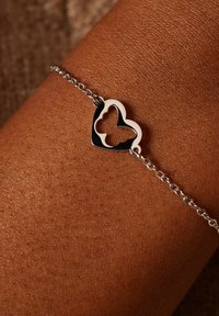 Zilveren kettingarmband met een hartvormige hanger met een vlinder uitsnede, uitgelegd tegen een gestructureerde huidachtergrond.
