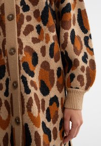 Beige kardigan s leopardím potiskem, se strukturovanými hnědými, oranžovými a černými skvrnami. Knoflíky vpředu a žebrované manžety.