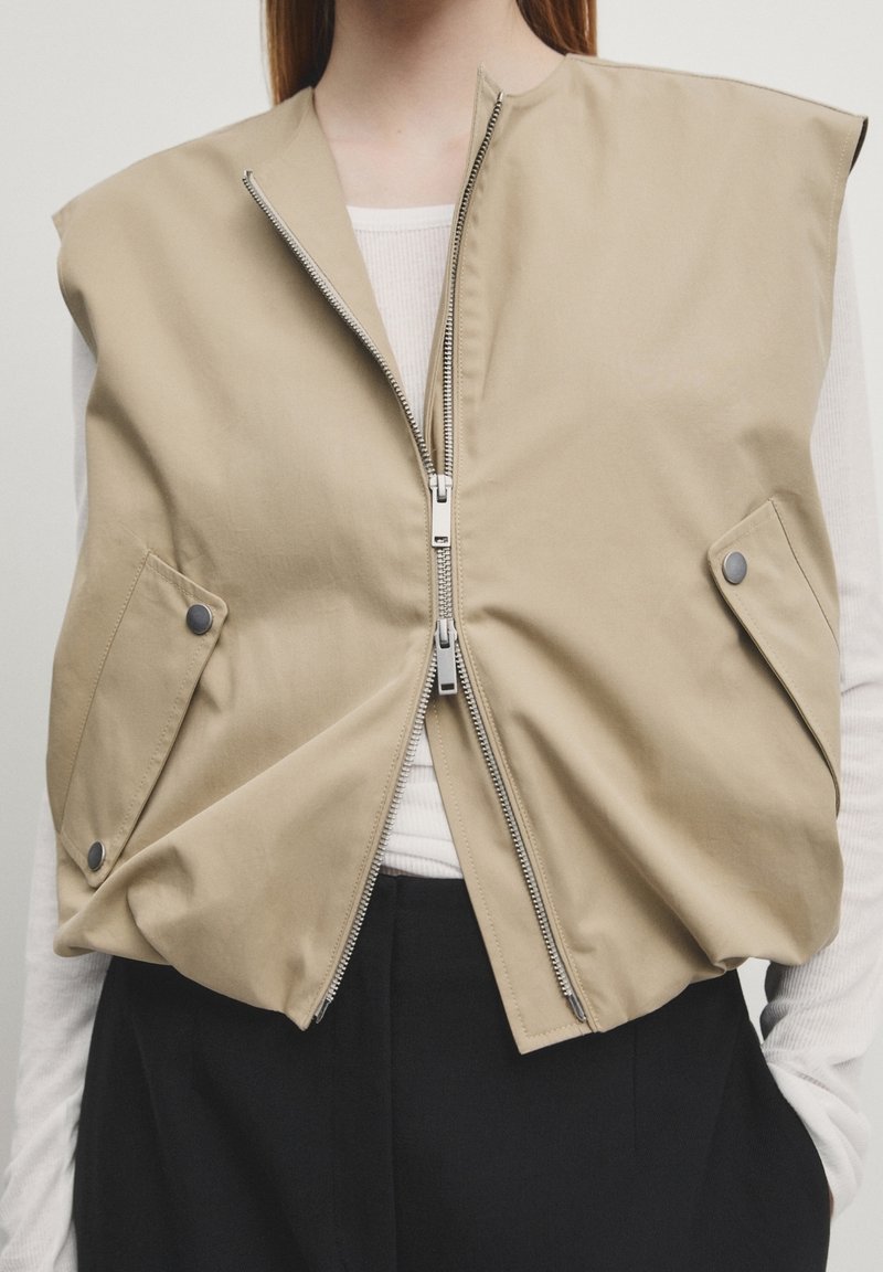 Veste beige oversize sans manches avec deux poches avant à pressions et une fermeture éclair double centrale portée sur un haut blanc à manches longues.