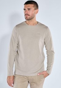 Beige Langarm-T-Shirt aus weichem Stoff, mit rundem Ausschnitt und einem dezenten, geprägten Logo auf der Brust.