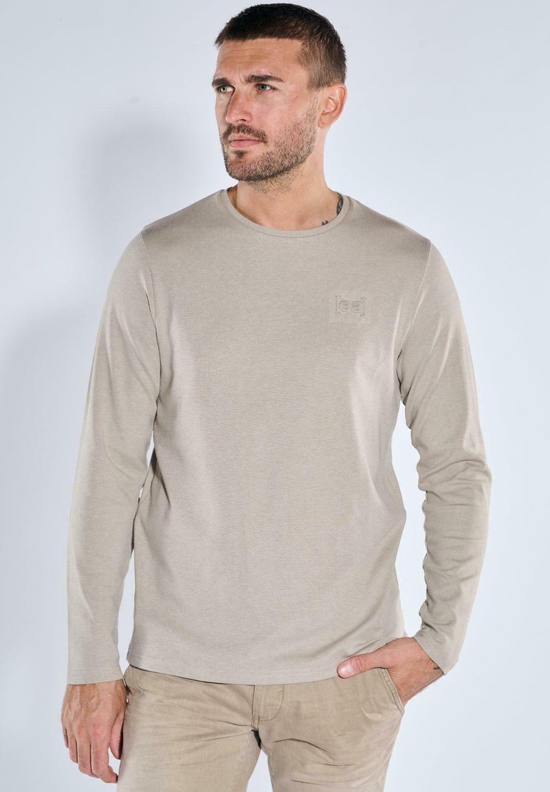 Beige Langarm-T-Shirt aus weichem Stoff, mit rundem Ausschnitt und einem dezenten, geprägten Logo auf der Brust.