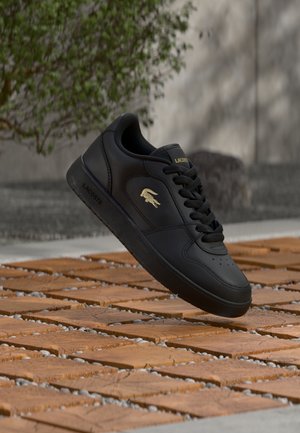 Zwarte Lacoste-sneaker met gouden logo, zwevend boven betegelde houten en kiezelsgrond, met een wazige achtergrond van groenblijvende planten en een grijze muur.