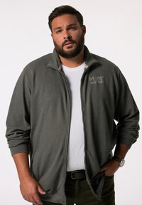 Chaqueta gris con cremallera en tejido texturizado y detalles en canalé, con bolsillos laterales y un pequeño logo en el pecho. Llevada sobre una camisa blanca.
