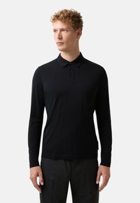 Mężczyzna z kręconymi blond włosami, ubrany w czarną koszulę polo z długim rękawem i ciemnoszare spodnie, spoglądający w lewo na jednolity szary tle.