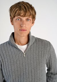 Szary sweter o prążkowanej fakturze, z wysokim kołnierzem i półzamkiem z przodu. Noszony na białej koszulce. Gładkie tło.