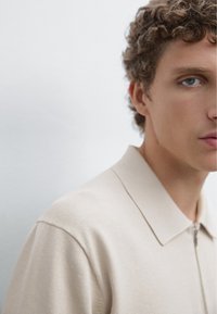 Beige pletená polo košile se zipovým zapínáním, širokým límcem a texturovanou tkaninou, částečně zobrazená na rameni a obličeji modelu.