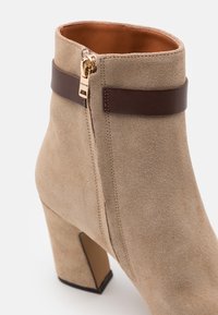 Botín de ante beige con tacón ancho, detalle de correa de cuero marrón y tirador de cremallera dorado grabado con "Coach."