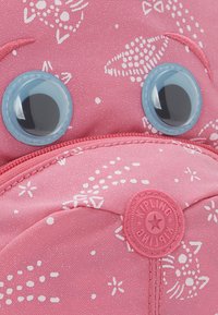 Rosa stof med hvide abstrakte kattemønstre, to store blå plastikøjne og et cirkulært Kipling-logo mærke.