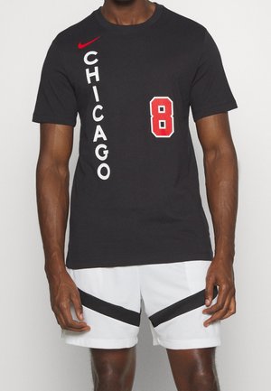 Homme portant un T-shirt noir avec le texte vertical "CHICAGO" et le chiffre rouge 8, associé à un short blanc avec des rayures diagonales noires.