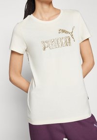 Kvinna iklädd en vit Puma-t-shirt med en gyllene zebrarandig logotyp och lila byxor med en liten vit Puma-logotyp.
