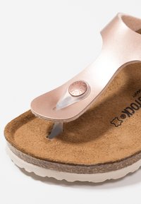 Infradito color oro rosa con una soletta in sughero texture, fascia trasparente e emblema rotondo in metallo. Design leggero con una finitura morbida.