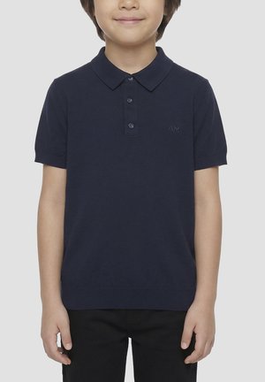 Kind draagt een navyblauw poloshirt met korte mouwen en drie knopen en een zwarte broek, staat met ontspannen armen langs het lichaam.