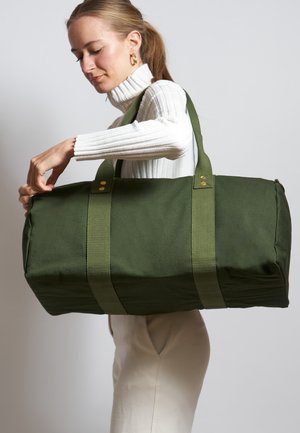 Sac de voyage en toile verte avec deux poignées supérieures, sécurisé par des éléments en métal doré, et une forme structurée, porté par une personne.
