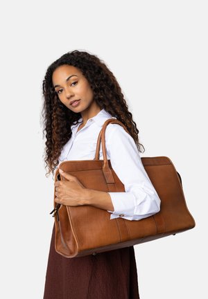 Donna con capelli ricci, che indossa una camicia bianca e una gonna marrone, mentre tiene una grande borsa a tracolla in pelle marrone.