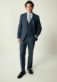Dunkelblauer Dreiteiler mit Revers, Knopfakzenten und einem hellblauen Hemd mit gemusterter Krawatte. Schwarze Schuhe sind sichtbar.