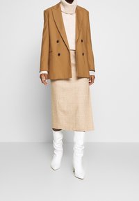 Brauner doppelt-knopfbarer Blazer über einem beigefarbenen Rollkragenpullover, kombiniert mit einem karierter Midirock und weißen kniehohen Stiefeln. Glatte Stoffe und strukturiertes Design.