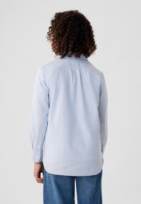 Asmuo su garbanotais plaukais, dėvintis šviesiai mėlyną ilgomis rankovėmis marškinius ir mėlynas džinsines kelnes, matomas iš nugaros priešais paprastą foną.