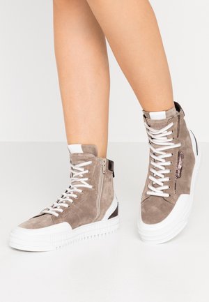 Nohy v taupových semišových vysokých teniskách s bílými tkaničkami, tlustými bílými podrážkami, bočními zipy a tmavými akcenty na patě.