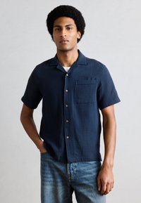 Un homme se tient debout, une main dans la poche, portant une chemise à manches courtes bleu marine boutonnée par-dessus un t-shirt blanc et un jean bleu délavé, devant un mur uni.