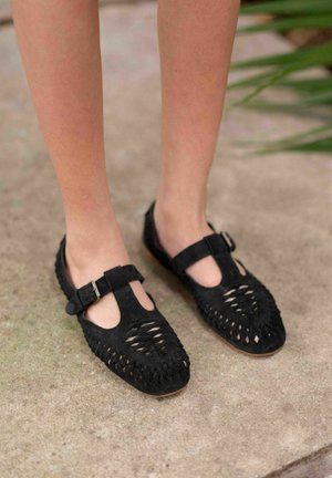 Ballerine nere in suede con un design a taglio, chiusura a fibbia con T-strap e cuciture testurizzate lungo la suola e la parte superiore. Suola piatta, senza tacco.