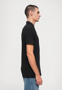 Svart polo skjorte i bomullsing, med korte ermer, med en klassisk krage og et ensfarget design. Modellen står sidelengs, iført lys blå jeans.