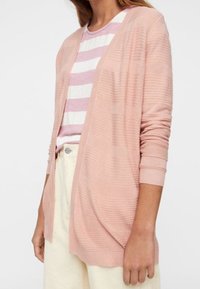 Jasnoróżowy sweter na drutach z prążkowaną teksturą i otwartym przodem. Noszony na fioletowo-białej koszuli w paski, z długimi rękawami.