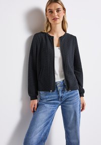 Kvinna som bär en svart texturerad zip-up jacka över en vit topp och ljusblå vida jeans, står mot en vit bakgrund.