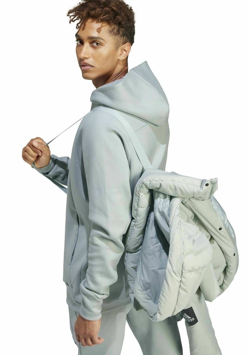 Jasnoszara bluza z kapturem, ze sznurkiem i kieszeniami, noszona z dopasowanymi spodniami. Osoba niesie jasnoszarą kurtkę puchową na jednym ramieniu.