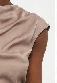 Haut en satin beige sans manches drapé sur l'épaule d'une personne à peau foncée, sur un fond blanc uni.