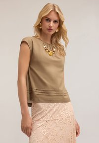 Beige short-sleeve top with pleated hem, paired with a light pink lace skirt, featuring intricate floral patterns. Gold statement necklace accentuates.