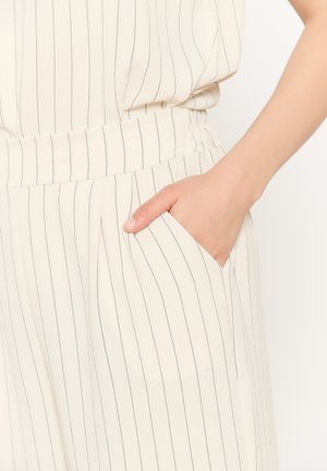 Ruka odpočívajúca vo vrecku bielych nohavíc s tenkými čiernymi zvislými pruhmi a zladeným pruhovaným topom.