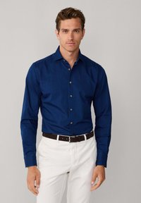Camicia blu navy con bottoni e colletto, con maniche lunghe e bottoni marroni, abbinata a pantaloni bianchi e cintura marrone scuro.