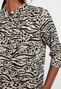 Černá a béžová košile na knoflíky s potiskem zvířecí kůže; má mandlový límec a vyhrnuté rukávy. Měkká látka s hladkou texturou.