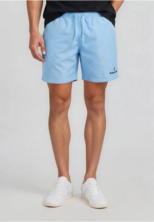 Mann trägt hellblaue Shorts mit Kordelzug und weiße Sneaker, steht vor einfachem Hintergrund.