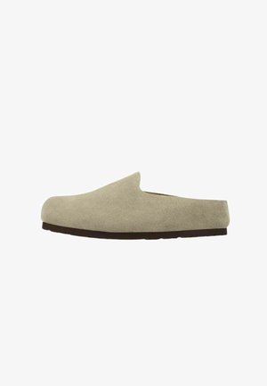 Scarpa mule beige in camoscio slip-on con tacco basso e suola piatta nera, vista di profilo su sfondo bianco.