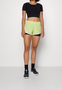 Svart kortärmad t-shirt, ljusgröna shorts med vit trim, lagda över svarta trosor och svarta träningsskor med vita sulor.