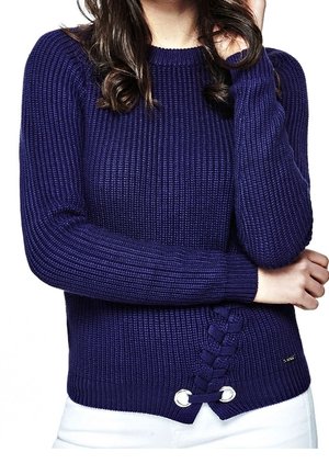 Žena v námořnicky modrém pleteném svetru s copánkovým detailem na lemu a bílých kalhotách, se zkříženýma rukama a jednou rukou blízko obličeje.