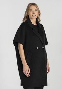 Czarna wełniana peleryna płaszcz z dwurzędowym zapięciem, kołnierzem z wycięciem, dwoma przednimi kieszeniami i krótkimi rękawami. Gładka tekstura.