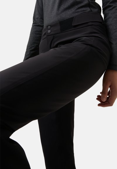 Sorte, tætsiddende bukser med en glat overflade, der har to knapper og en velcro-lukning i taljen. Sidelommer er synlige.