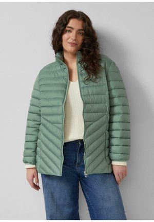 Kobieta o długich kręconych włosach, ubrana w jasnozieloną pikowaną kurtkę na kremowym swetrze i niebieskich dżinsach, stoi na szarym tle.