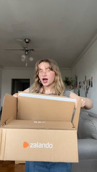 Cardboard box with Zalando logo, opened wide, held by a person. Gray walls and light-colored furniture visible in background.