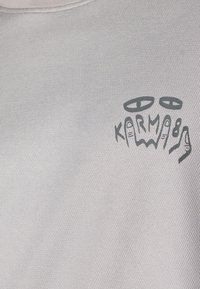 T-shirt en tissu texturé respirant de couleur gris clair avec un logo graphique noir "KARMA" présentant des mains stylisées et des yeux.
