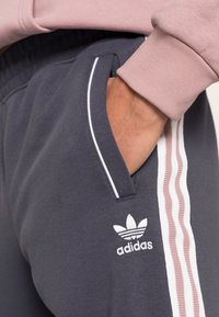 Pantaloni tuta grigi con strisce laterali bianche e rosa, dotati di tasche e con il logo Adidas ricamato in bianco sulla gamba inferiore.