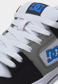 Άσπρο, γκρι και μαύρο αθλητικό παπούτσι με άσπρα κορδόνια και μπλε λογότυπο DC στο πλάι και στη γλώσσα, σε κοντινή λήψη.