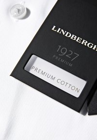 Camicia bianca con un motivo testurizzato, presenta un bottone rotondo bianco e un'etichetta nera con la scritta "LINDENBERGH 1927 PREMIUM COTTON."